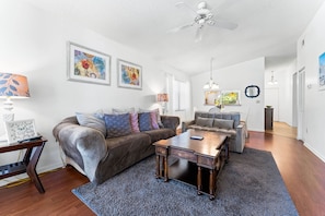 A living room with a brown sofa, a wooden coffee table, and a gray armchair. There are two framed artworks on the wall, a ceiling fan, and a chandelier.