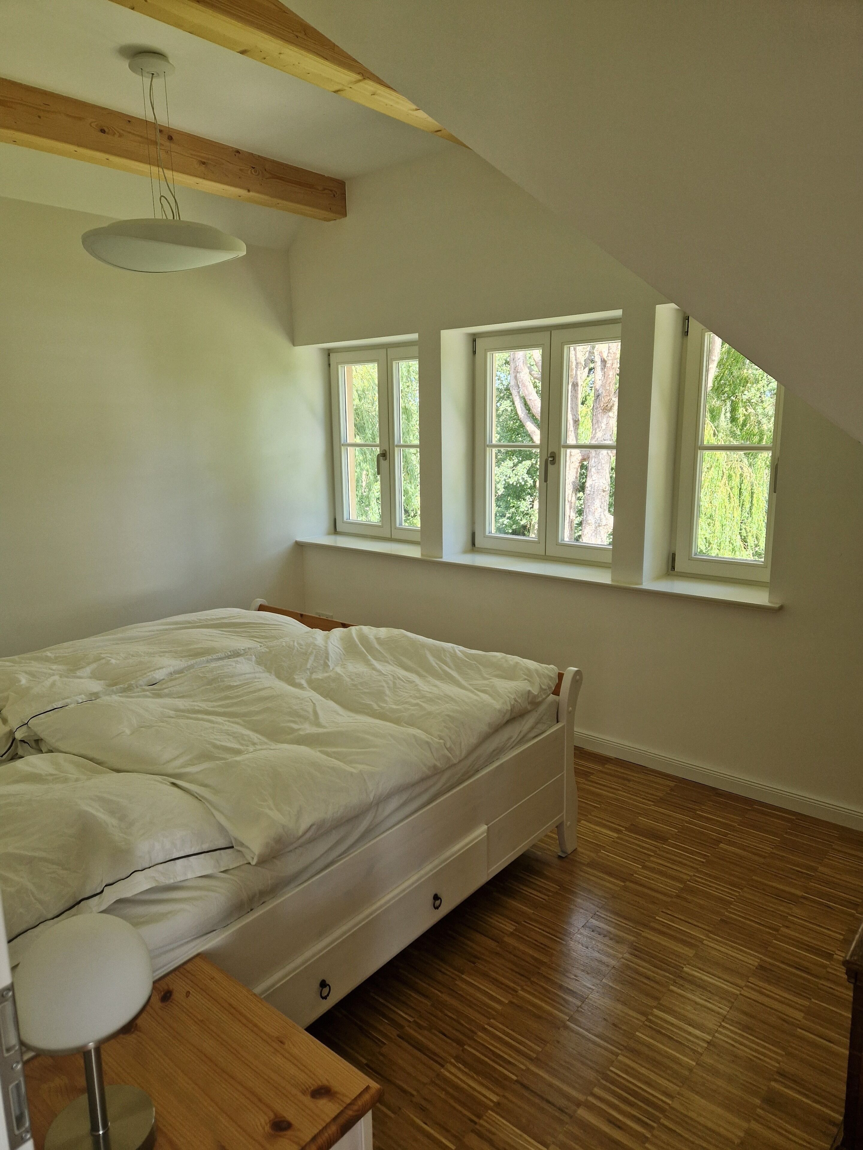 Une chambre avec un plancher en bois, un lit avec une literie blanche, une table de chevet et une fenêtre donnant sur des arbres.