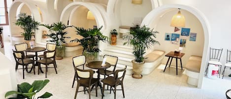 Un interior moderno con mesas y sillas redondas, plantas en macetas y una escalera con un banco color beige.