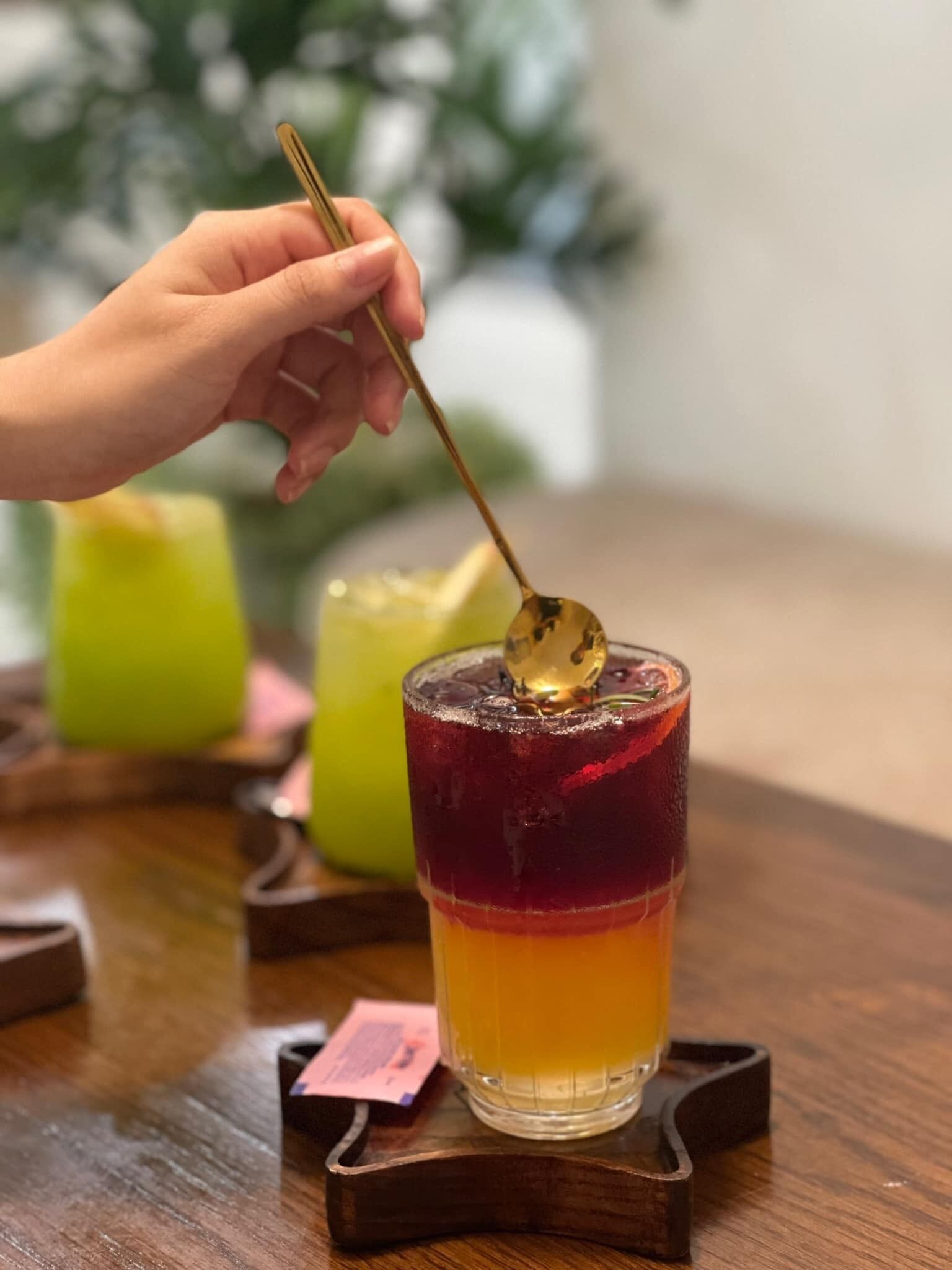 Une main tenant une cuillère au-dessus d’une boisson à plusieurs couches, servie dans un verre posé sur un sous-verre en bois.