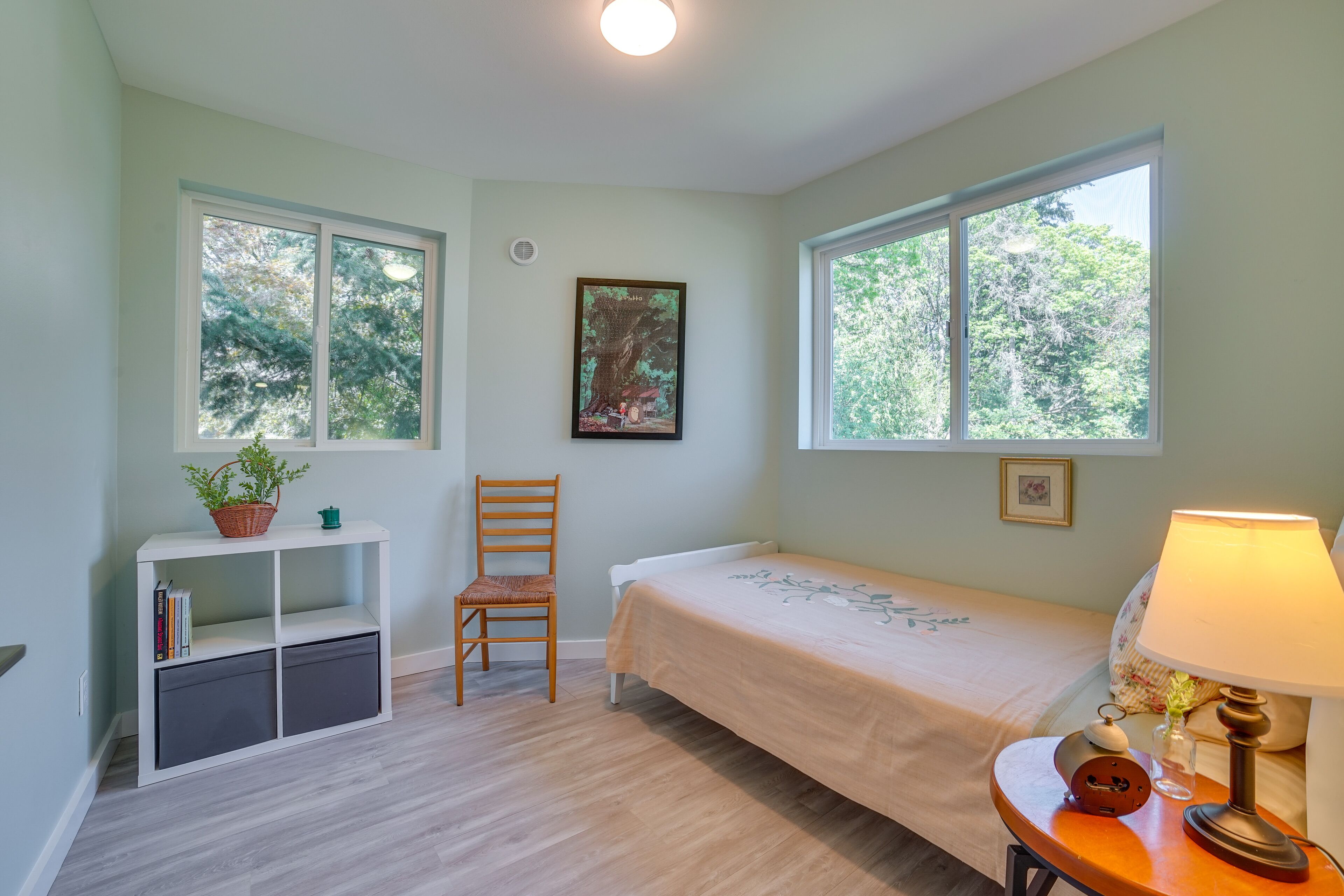 A room with a bed, a chair, a small table with a lamp, a bookshelf, and two windows with a view of trees.
