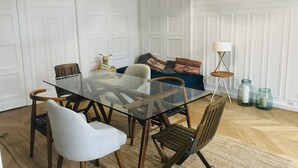 A dining area with a glass table, wooden chairs, a sofa with patterned cushions, and a floor lamp.