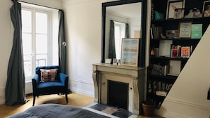 A bedroom with a large bed, a blue armchair, a fireplace, a bookshelf, and a large mirror.