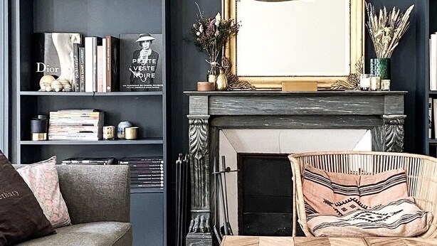 A living room with a dark blue wall, a fireplace, a wooden coffee table, a grey sofa, and a bookshelf filled with books and decorative items.