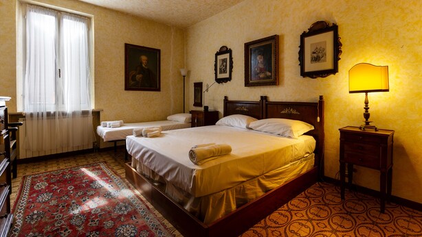 A hotel room with a large bed, two smaller beds, a wooden headboard, a nightstand with a lamp, and a patterned carpet. - Apartment in the historic center with private car parking (Mantova)