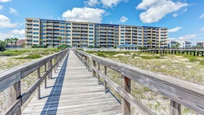 Een houten looppad leidt naar een meerlaags hotel met balkons, gelegen aan een strand met weinig begroeiing.