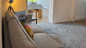 En moderne stue med en beige sofa, en lampe og udsigt til et spiseområde gennem en glasdør.