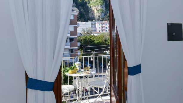 A balcony with white curtains, a table with chairs, and a view of buildings on a hillside. - Natalia House center of Sorrento (Sorrento)
