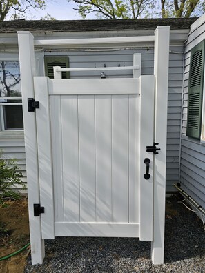 A white wooden gate with black hardware, attached to a house with white siding and green shutters. - Newly Renovated Mid-Cape Home with Pond and Waterfront Access (Dennis)
