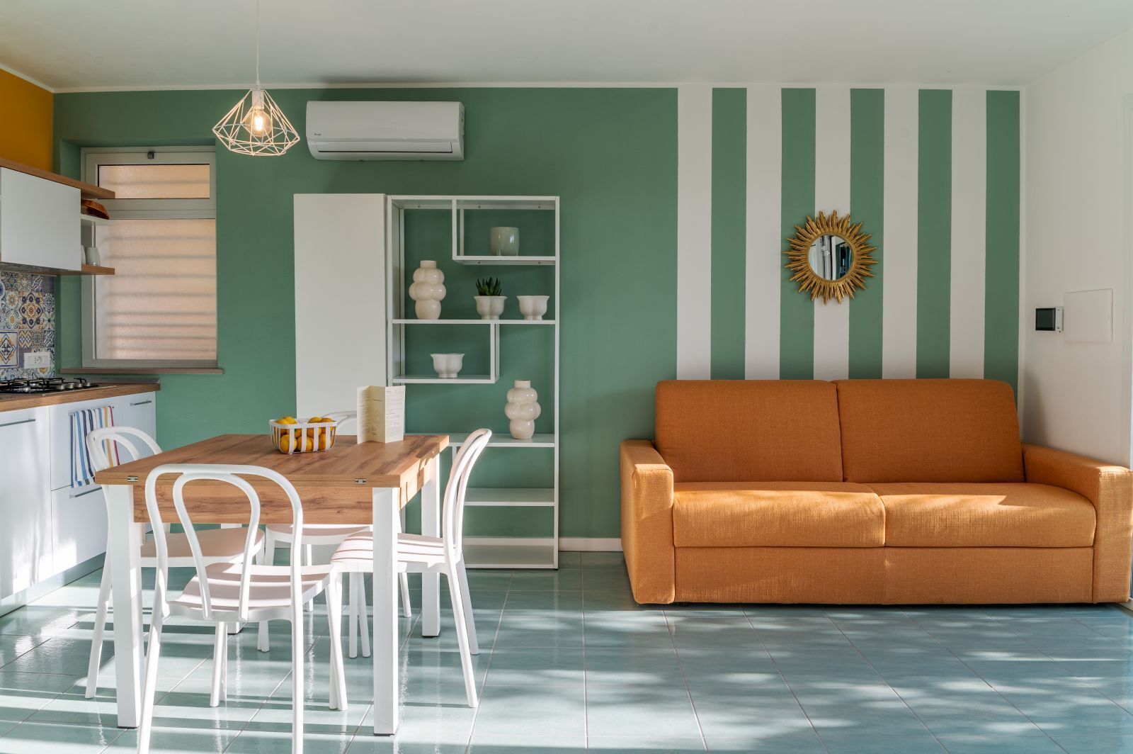Une cuisine moderne avec des meubles blancs, une table à manger en bois et des chaises blanches. Un mur à rayures vertes est agrémenté d’un miroir décoratif en forme de soleil, et une étagère blanche expose divers objets décoratifs.