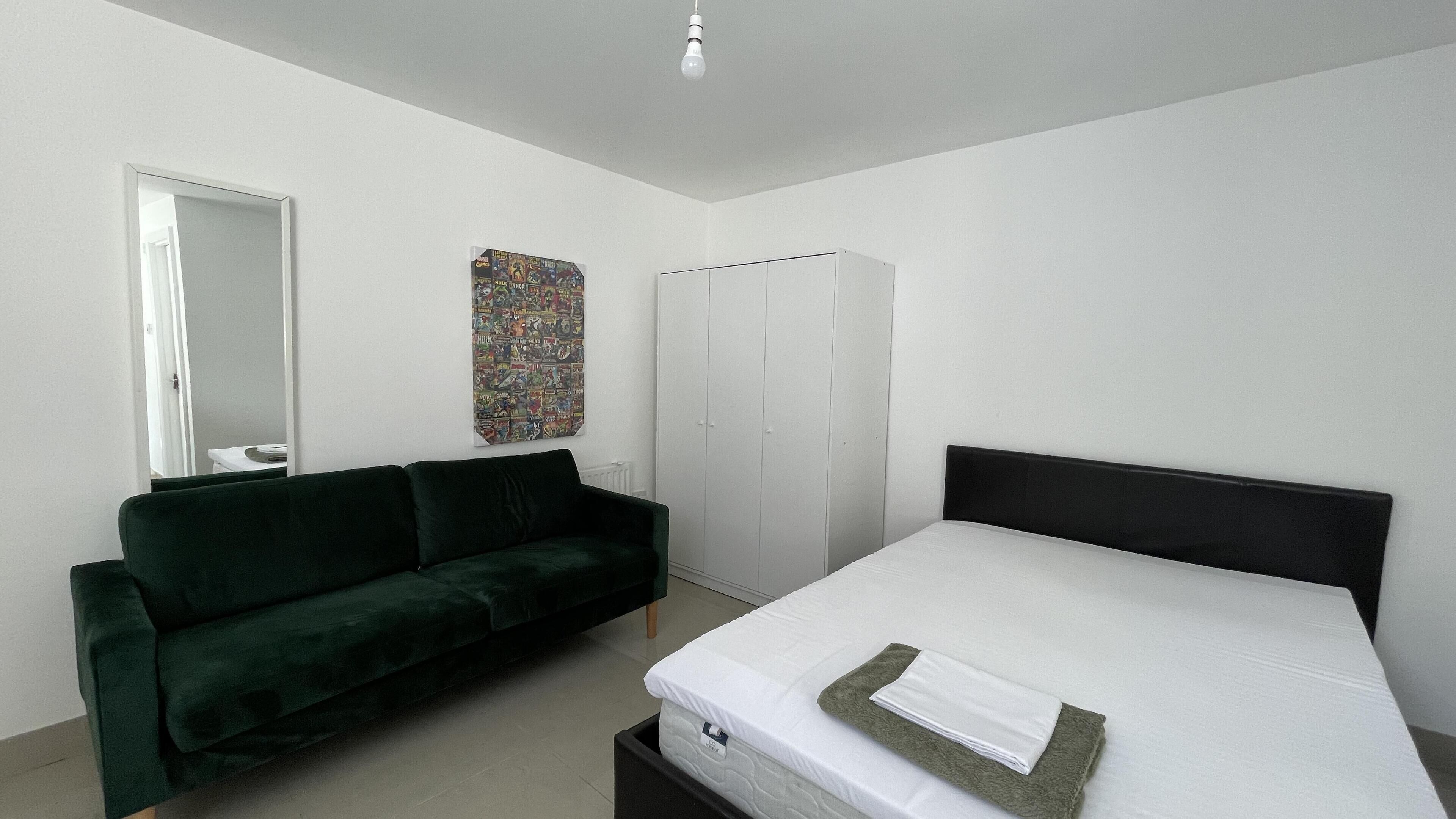 Un dormitorio con cama, un sofá verde oscuro, un armario, un espejo y un póster colorido en la pared.