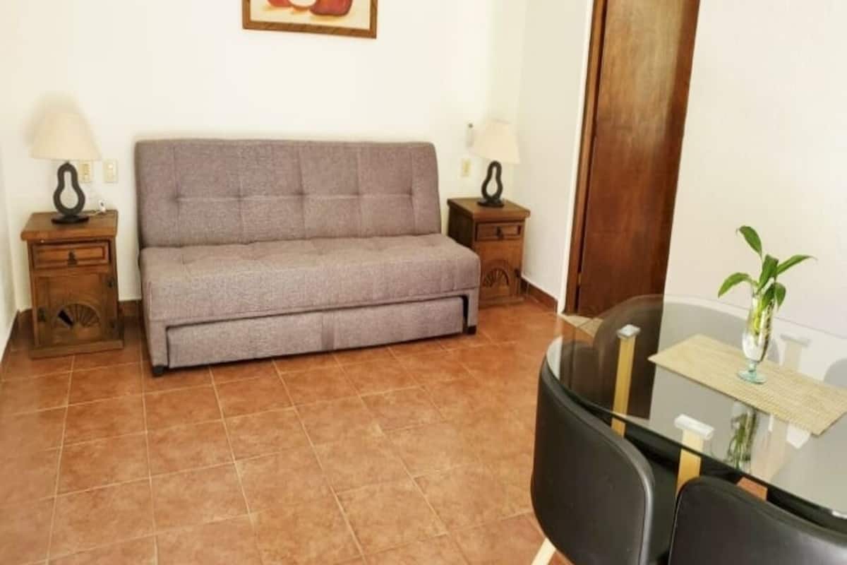 A living room with a sofa, two side tables with lamps, a glass table with chairs, and a potted plant.