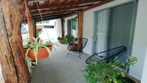 Un espacio exterior techado con plantas en macetas, dos sillas y un ventanal corredera.