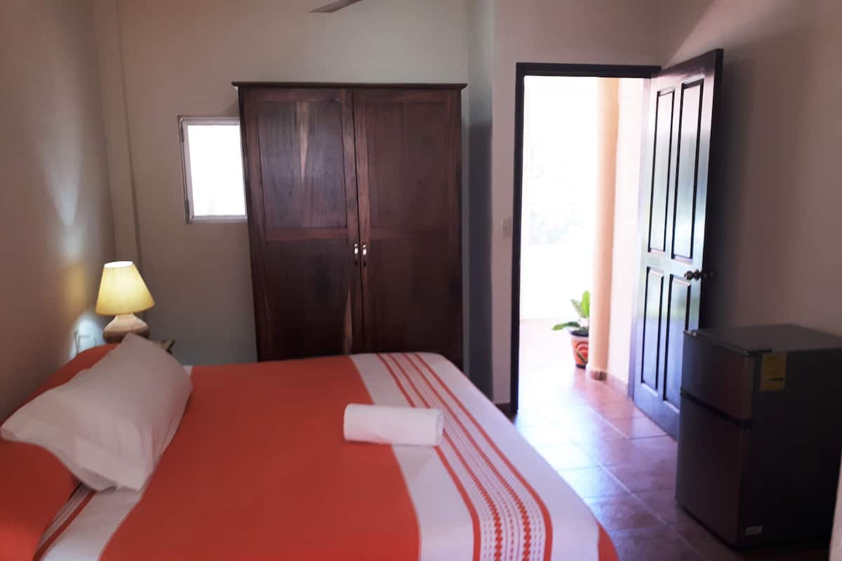 A bedroom with a bed, a wardrobe, a desk with a lamp, and a refrigerator.