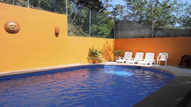 Una piscina con borde curvo, rodeada por un muro amarillo y una valla. Hay sillas reclinables blancas a lo largo del muro.