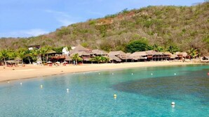 Una playa tropical despejada con costa rocosa, una playa de arena y cabañas con techo de paja al fondo.