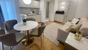 Una sala de estar moderna con una mesa de comedor redonda, sillas grises, un sofá con cojines, una máquina de café y una zona de cocina con un microondas.