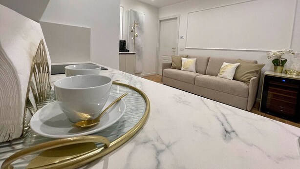 Una sala de estar moderna con encimera de mármol, un sofá y un comedor con mesa de vidrio y platos con detalles dorados.