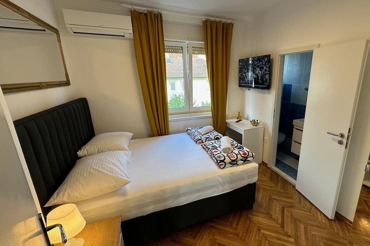 A hotel room with a bed, a bedside lamp, a window with curtains, a TV, and a bathroom visible through an open door.