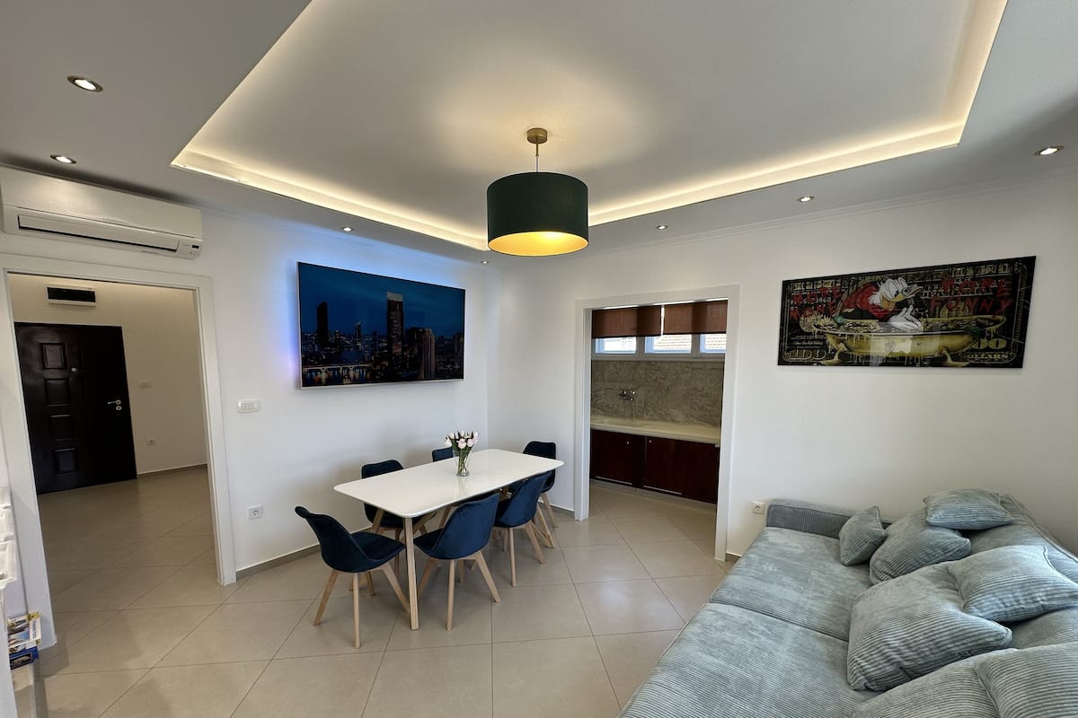 A modern living room with a sofa, dining table, and chairs. There is a kitchen area with a sink and cabinets.