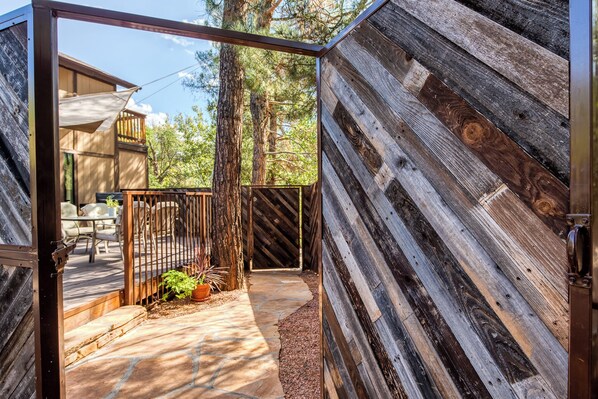 A wooden deck with a stone pathway, a fenced area with a table and chairs, and a view of trees.