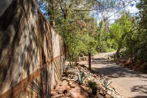 A paved pathway flanked by a wooden fence and various plants, leading through a wooded area.