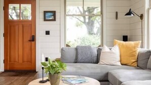 A living room with a grey sofa, a wooden door, and a window with a view of greenery.
