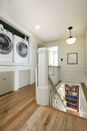 Un cuarto de lavandería con lavadoras y secadoras apiladas, una escalera que baja a un nivel inferior y una estantería con libros.