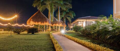 A pathway leading to a building with a thatched-roof structure, surrounded by well-maintained grass and palm trees.