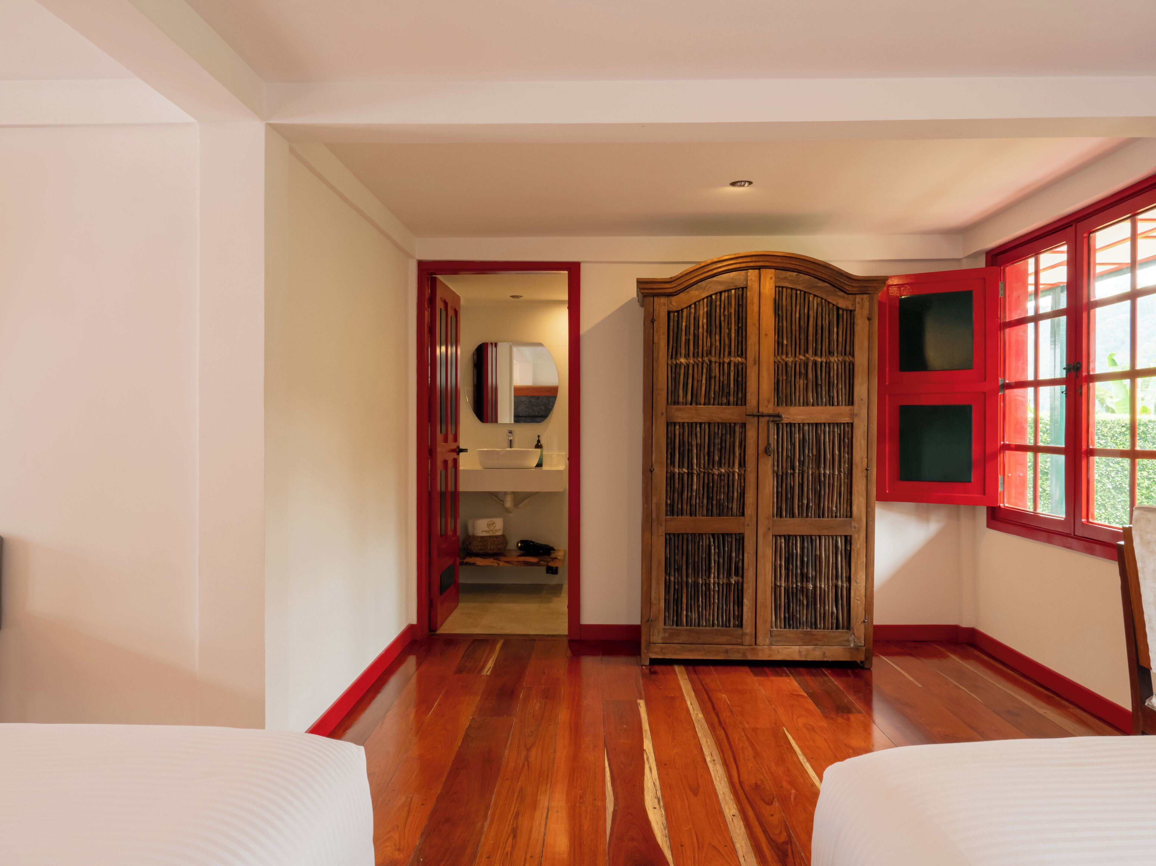 A room with wooden flooring, a wardrobe with a mirror, and a window with red shutters.