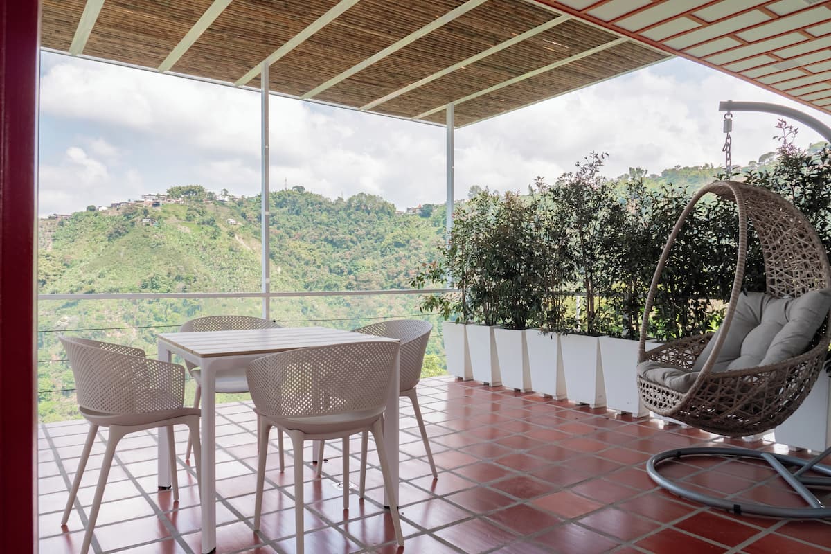 A balcony with a table and chairs, a hanging chair, and a view of a green hillside.