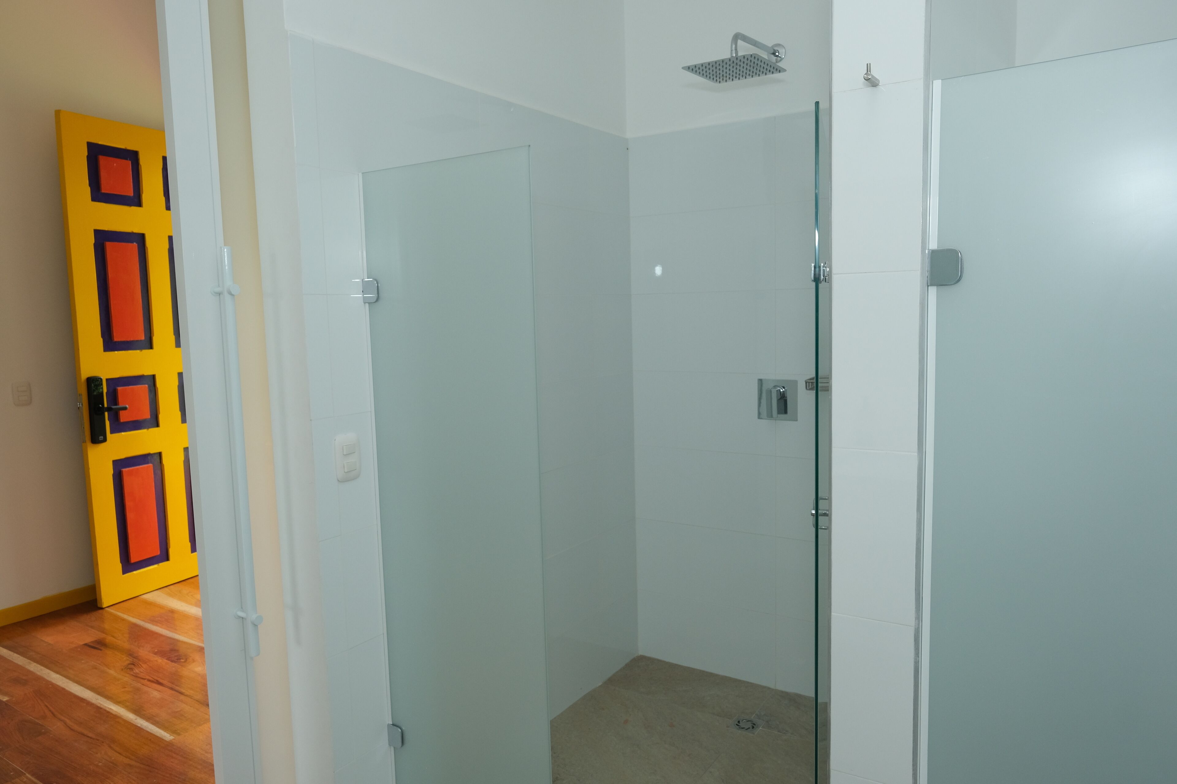 A bathroom with a glass shower enclosure, white tiled walls, and a wooden floor.
