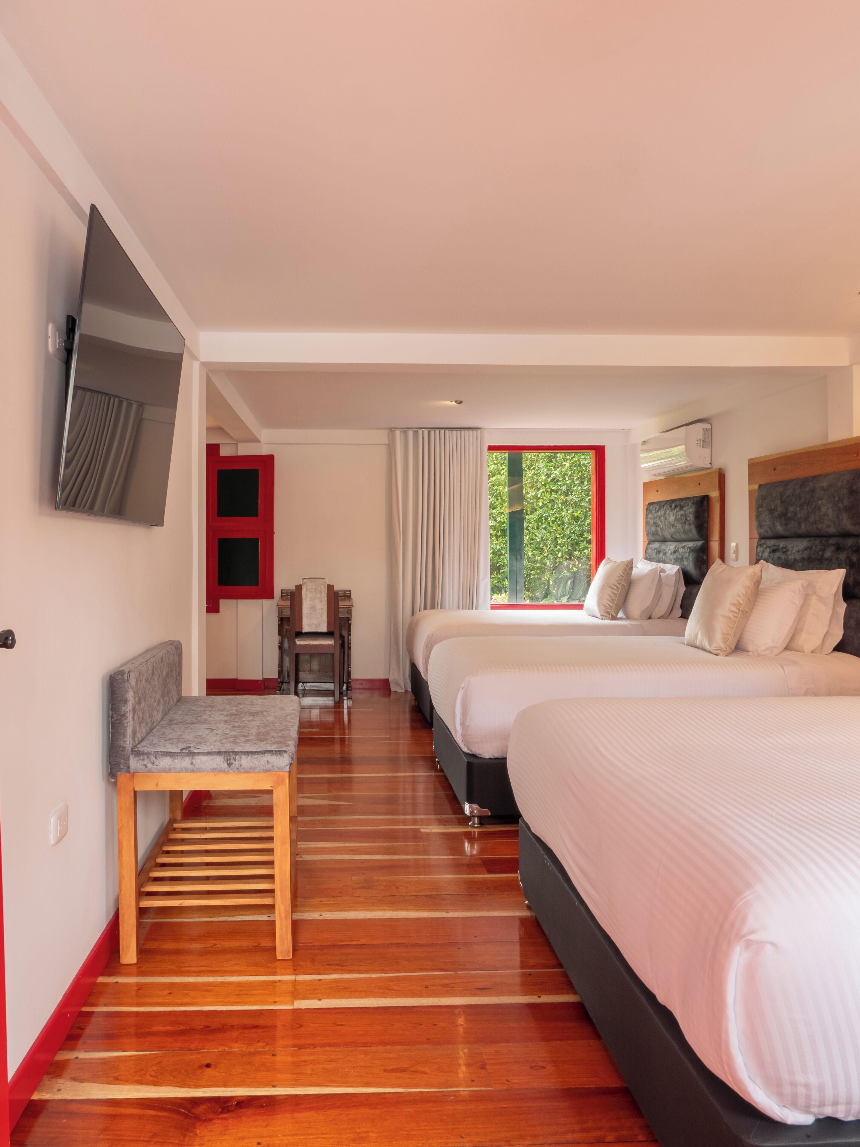 Habitación de hotel con dos camas, un televisor montado en la pared, un banco de madera y una ventana con vistas al verdor.