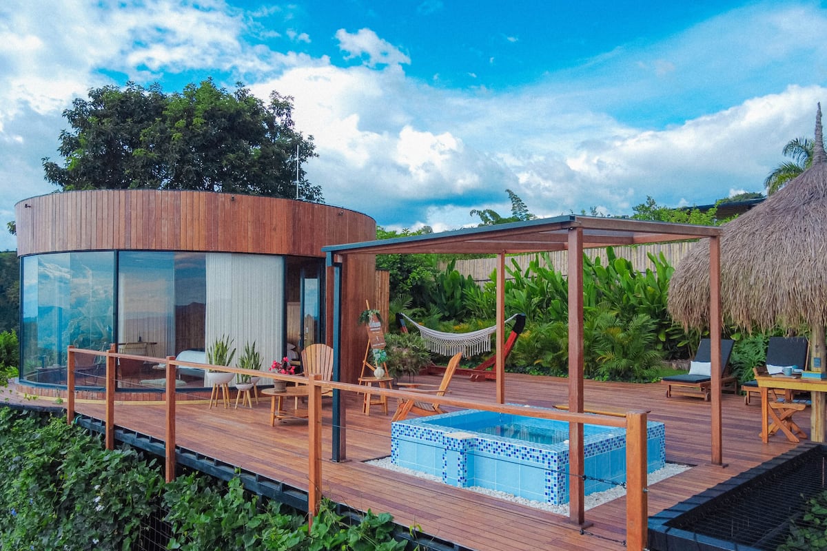 A modern outdoor deck with a glass-enclosed room, a wooden deck, lounge chairs, a hammock, and a hot tub.