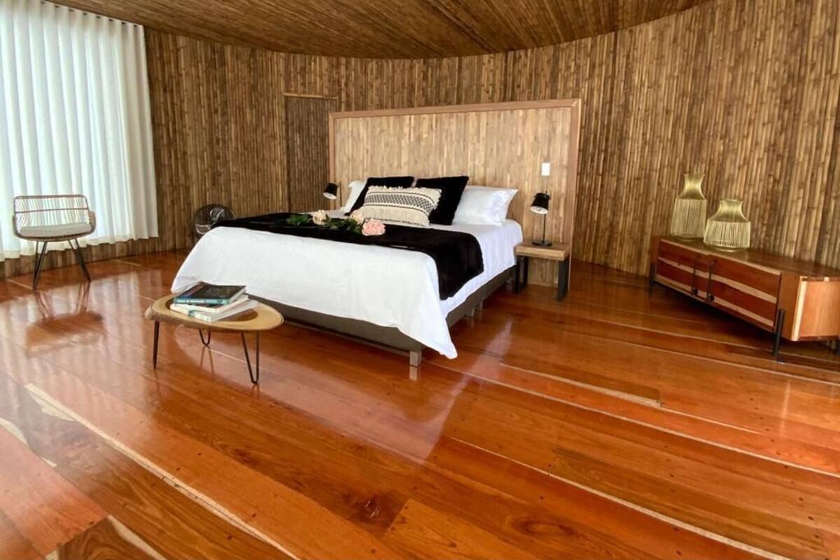 A room with a wooden floor, a bed with white bedding, a bedside table with books, and a chair.