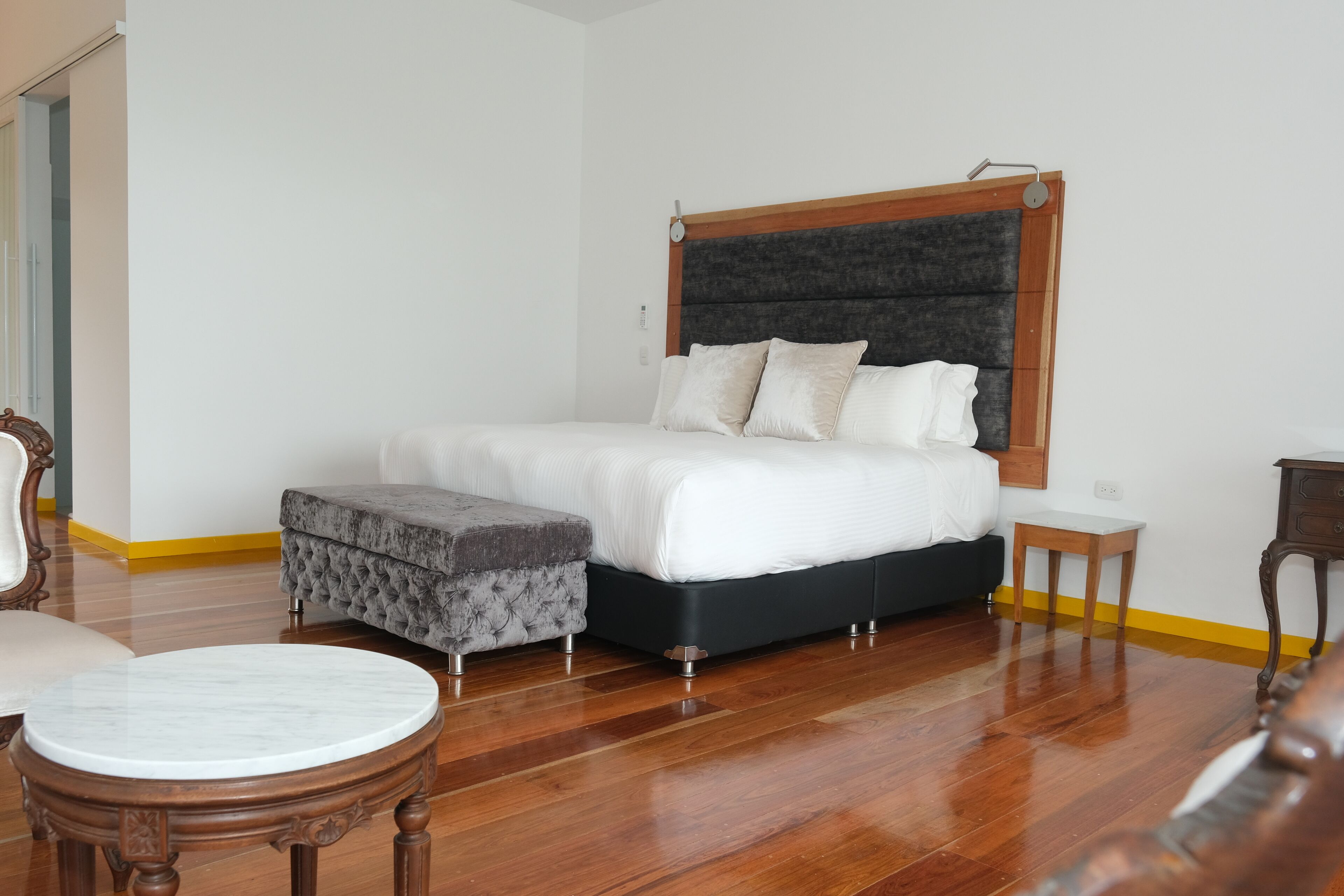 Un dormitorio con una cama grande, un cabecero de madera, una mesita de noche pequeña, un otoman tapizado y una mesa redonda de madera.