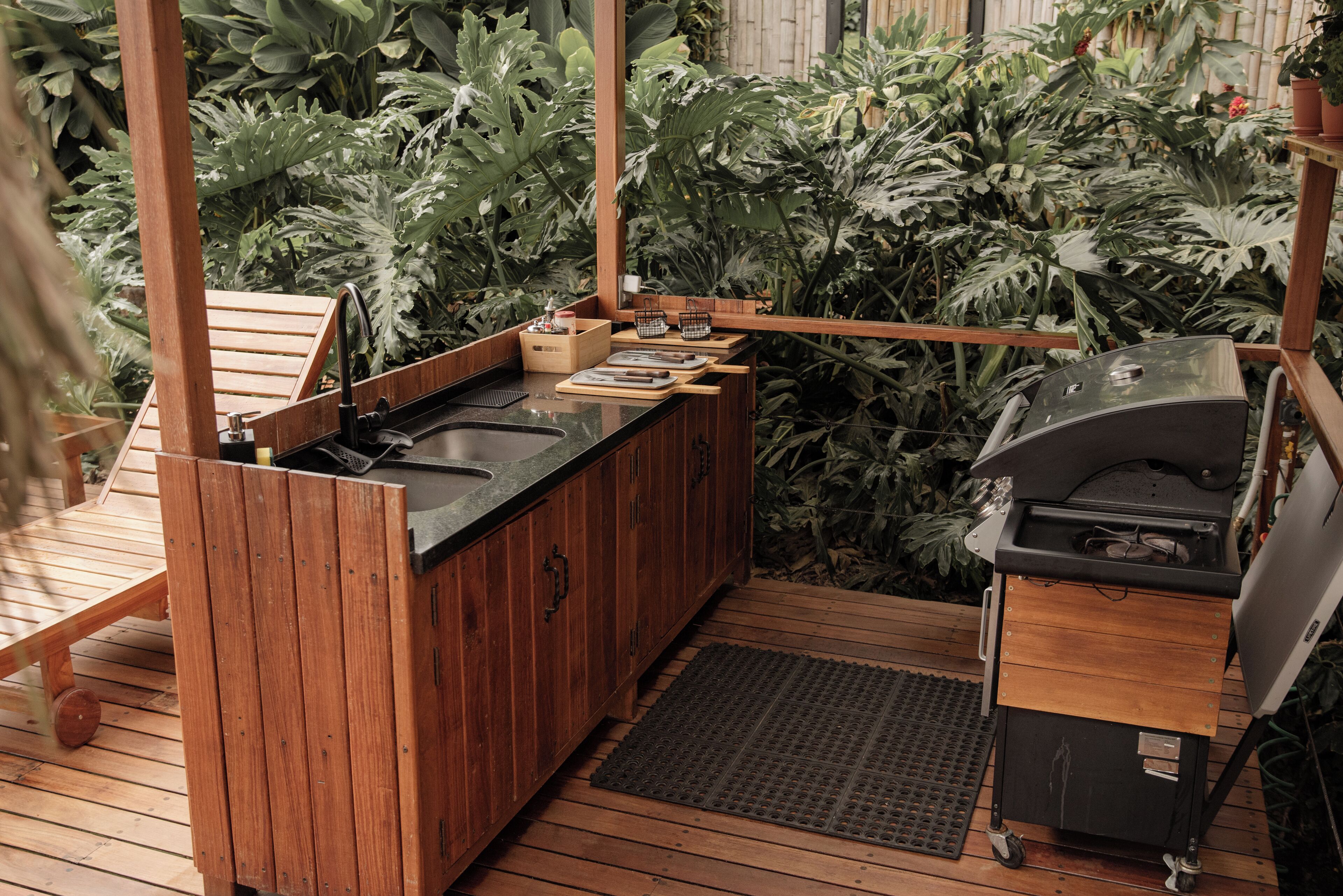 Une cuisine extérieure en bois, dotée d’un évier, d’un plan de travail et d’un barbecue, le tout entouré d’une végétation luxuriante.