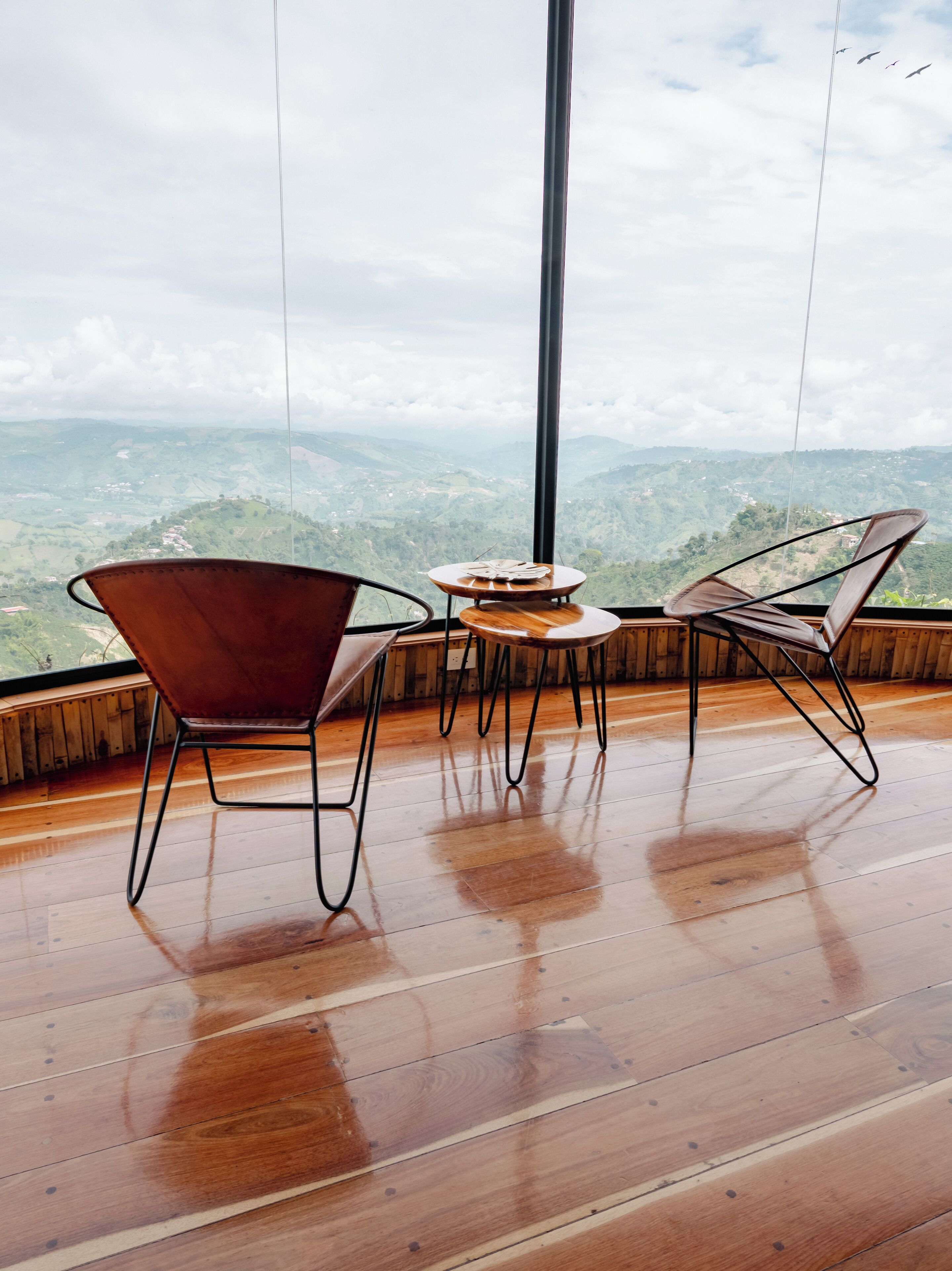 Une pièce dotée d’un plancher en bois, de deux fauteuils marron, d’une petite table ronde et d’une grande fenêtre donnant sur un paysage montagneux.