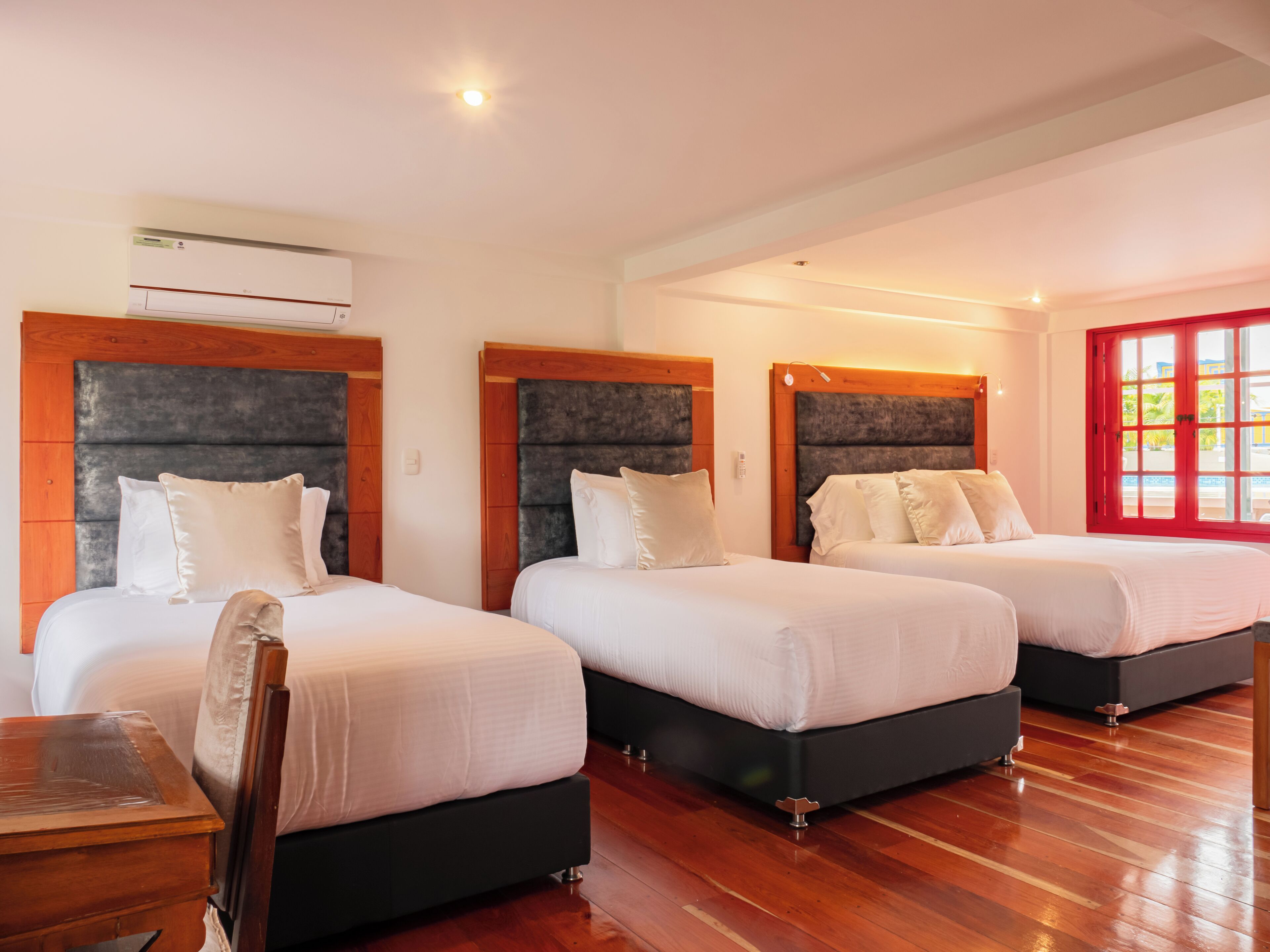 Een hotelkamer met drie bedden, houten vloeren en een raam met rode lijst.