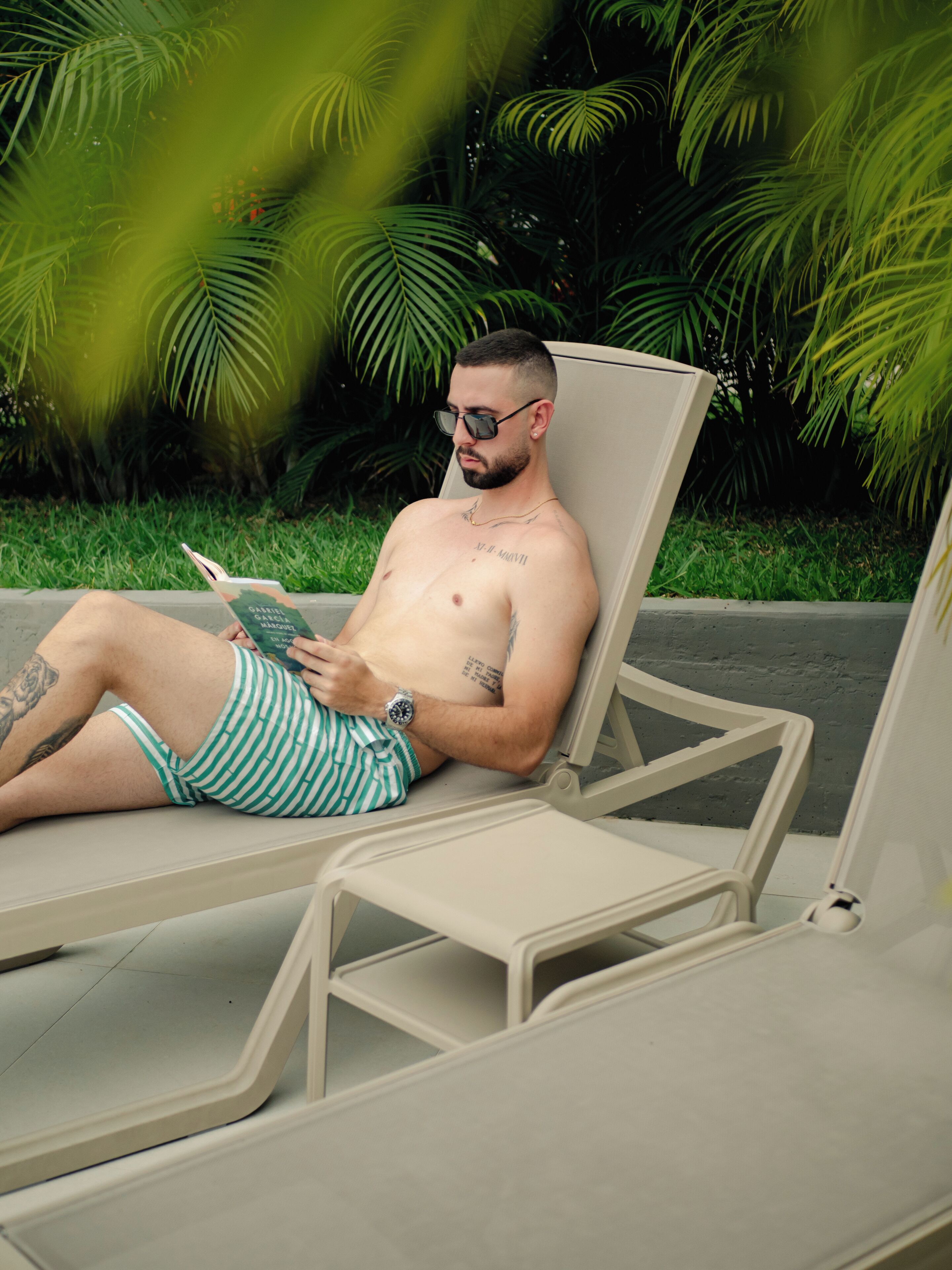 A man lying on a sun lounger reading a book, with a tattoo on his arm and a watch on his wrist.