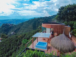 Une villa moderne dotée d’une piscine à débordement entièrement vitrée, d’une terrasse en bois et d’une structure à toit de chaume.