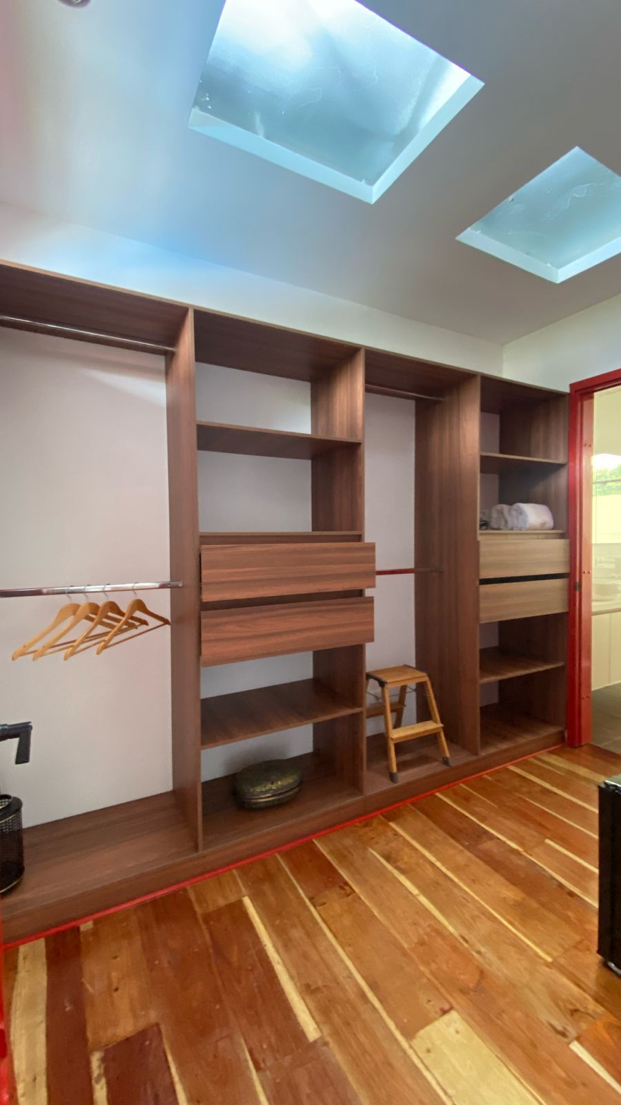 A room with a wooden floor, a built-in wardrobe with multiple shelves, a small stool, and a skylight.