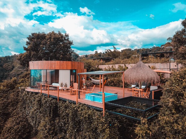 Une villa moderne dotée d’un salon entièrement vitré, d’une terrasse en bois et d’une structure surmontée d’un toit de chaume.