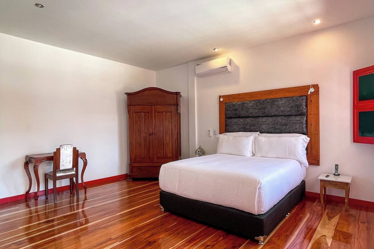 A bedroom with a wooden floor, a bed with white bedding, a wooden wardrobe, a small table, and a chair.