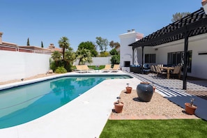 A swimming pool with a white deck, surrounded by a white wall and palm trees. There is a covered patio area with outdoor furniture. - Sunshine Villa by HomeSlice Stays (Phoenix)