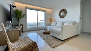 Un salon moderne avec un canapé blanc, un fauteuil en osier, une table en verre ornée d’une plante et une grande fenêtre offrant une vue dégagée.