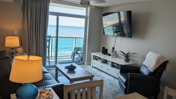 A living room with a sofa, a coffee table, a TV mounted on the wall, and a view of the ocean through a large window.