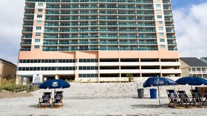 A multi-story hotel with a beach access sign, outdoor seating, and a parking structure.