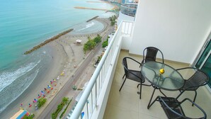 Ein Balkon mit Glastisch und Stühlen mit Blick auf einen Strand mit Sonnenschirmen und Personen.