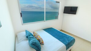 Ein Einzelbett mit Blick auf den Ozean durch ein großes Fenster, ein an der Wand befestigter Flachbildfernseher und ein gefliester Boden.